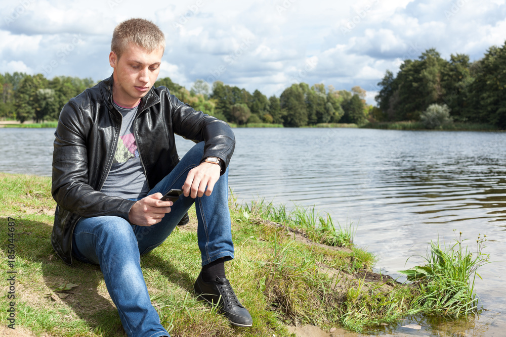 Blond man in black leather jacket sitting on river bank with mobile phone in hand
