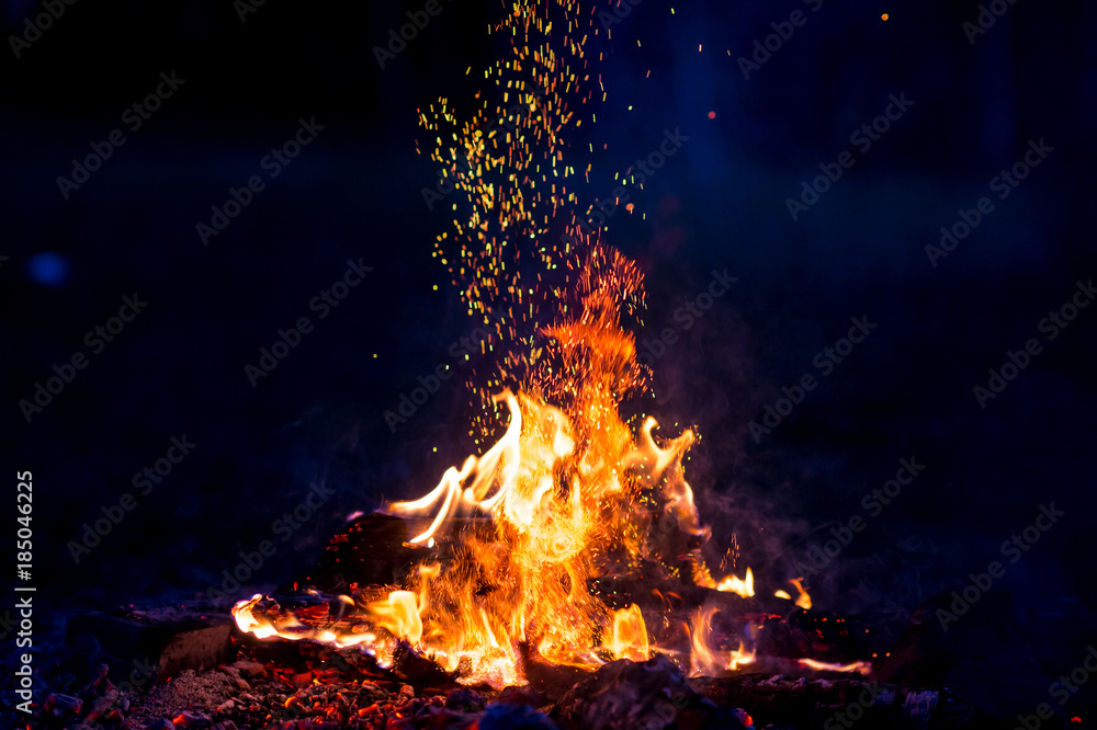 Burning wood at night. Campfire at touristic camp at nature in mountains. Flame amd fire sparks on dark abstract background. Cooking barbecue outdoor. Hellish fire element. Fuel, power and energy.
