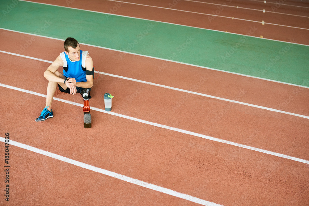 High angle portrait of young amputee athlete sitting on running track taking break from practice to relax, copy space High angle portrait of young amputee athlete sitting on running track taking break from practice to relax, copy space