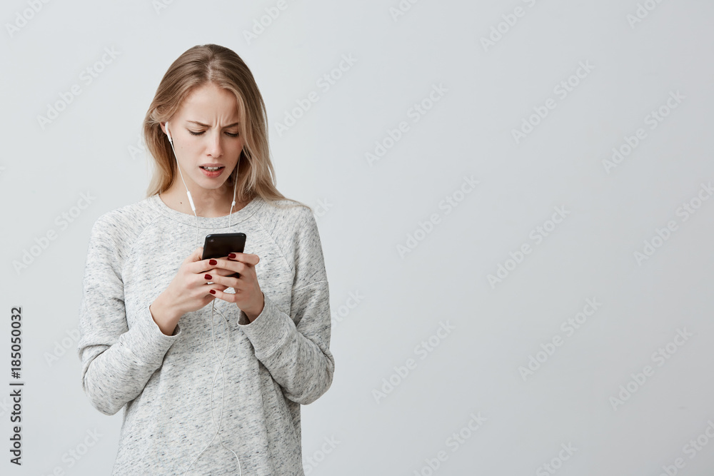 Amazed dissatisfied woman with dyed blonde hair dressed casually with white earphones holding smartphone recieving message being shocked to forget about important meeting with businesspartners Amazed dissatisfied woman with dyed blonde hair dressed casually with white earphones holding smartphone recieving message being shocked to forget about important meeting with businesspartners