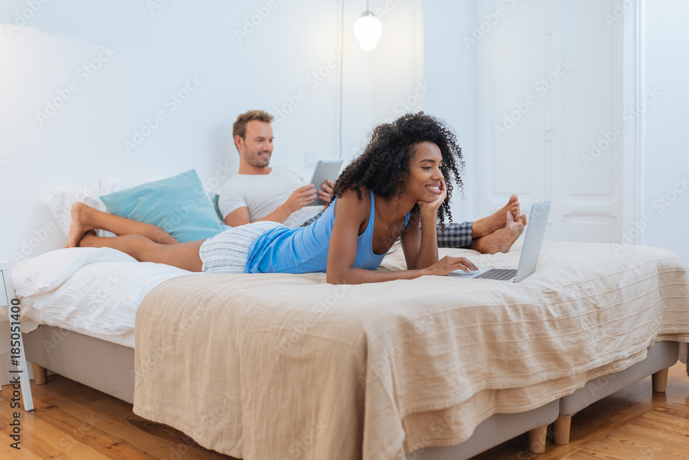 Happy young couple relaxed at home lying down in bed on the tablet and computer Happy young couple relaxed at home lying down in bed on the tablet and computer