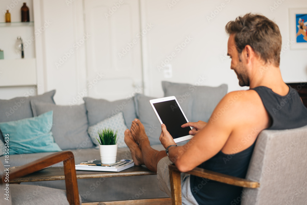 Happy young blonde man sitting in the couch at home on the tablet . Happy young blonde man sitting in the couch at home on the tablet .