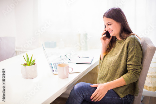 Side view of the young woman who sitting at the table and looking at laptop and speaking by phone and smiling at home