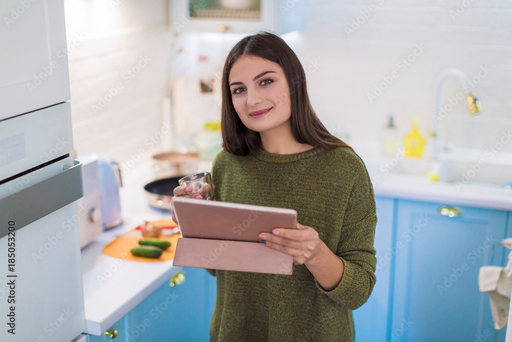 Portrait of the young smiling woman who standing in the kitchen and holding the tablet and glass in hands Portrait of the young smiling woman who standing in the kitchen and holding the tablet and glass in hands