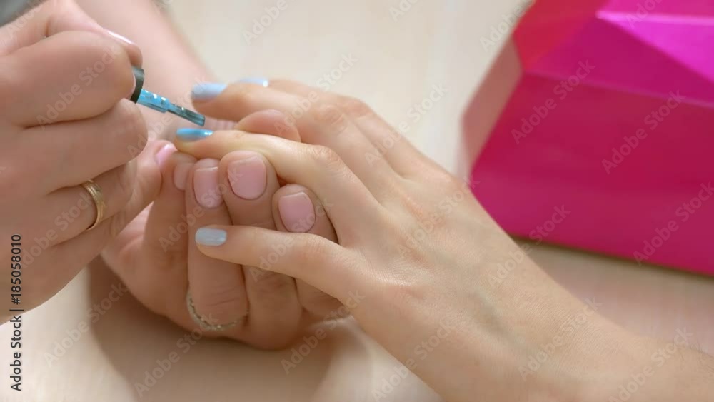 Manicurist covering nail with varnish close up. Manicure specialist applying blue color polish on client nail. Desktop with ultraviolet lamp and manicure process. Manicurist covering nail with varnish close up. Manicure specialist applying blue color polish on client nail. Desktop with ultraviolet lamp and manicure process.
