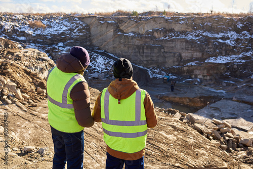 Back view portrait of two industrial workers wearing reflective jackets, one of them African, standing on cliff overlooking mineral mines on worksite outdoors, copy space