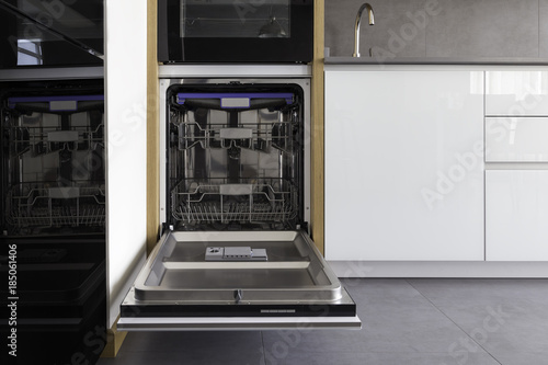 Opened new empty dishwasher in a modern loft kitchen in white, grey colors and wooden details. Incorporated dishwasher in the cabinets with hidden profile handle. 