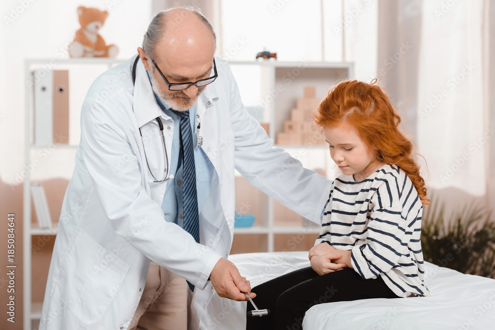 senior pediatrician in white coat examining little patient with reflex hammer in clinic senior pediatrician in white coat examining little patient with reflex hammer in clinic