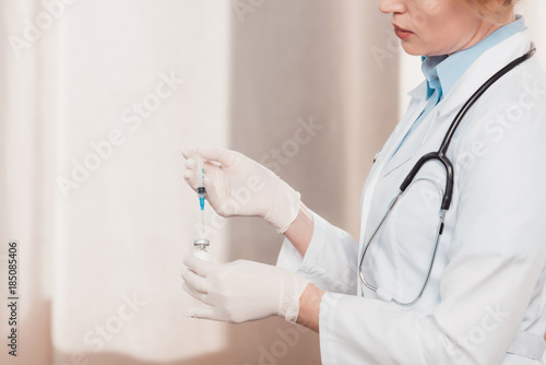 partial view of doctor in white coat and medical gloves holding syringe for injection in hands in clinic