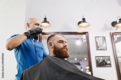 Male professional hairdresser serving client by scissors. Ginger handsome brutal stylish young man with thick big beard, short hair getting trendy haircut in black cape. Light white barber shop room.