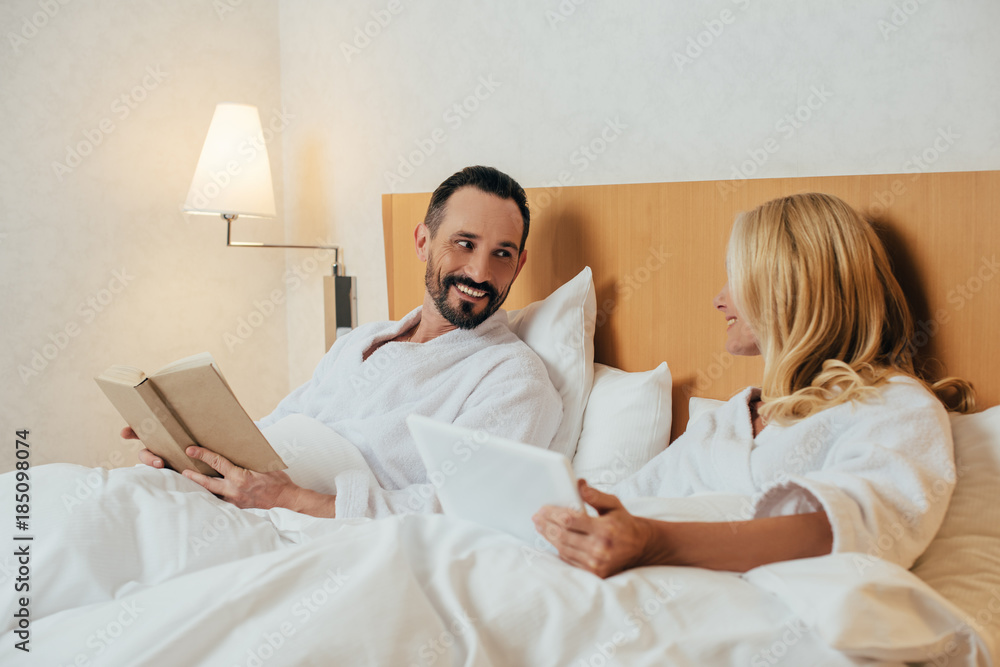 middle aged couple with digital tablet and book smiling each other while lying in bed at hotel room middle aged couple with digital tablet and book smiling each other while lying in bed at hotel room