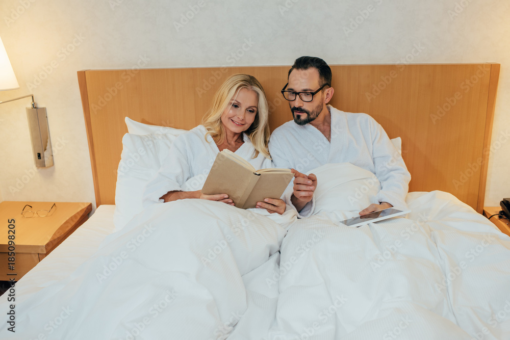 mature couple in bathrobes reading book and using digital tablet in bed in hotel room mature couple in bathrobes reading book and using digital tablet in bed in hotel room