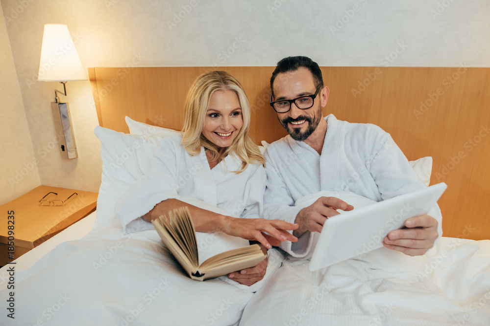 smiling mature couple reading book and using digital tablet while lying together in bed in hotel room smiling mature couple reading book and using digital tablet while lying together in bed in hotel room