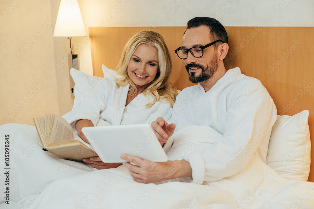 happy middle aged couple reading book and using digital tablet while lying together in bed in hotel room happy middle aged couple reading book and using digital tablet while lying together in bed in hotel room