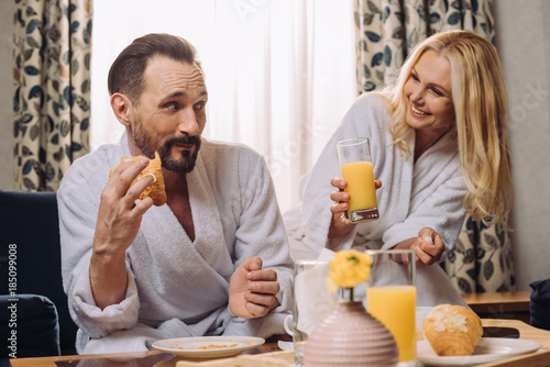 smiling middle aged couple drinking juice and eating pastry during breakfast in hotel room