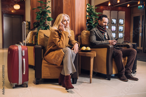 middle aged couple using smartphones while waiting with suitcases in hotel