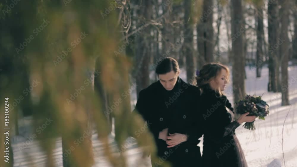 unusual wedding happy young lovers in black stand under tall pine tree. Gotic style. Beautiful brunette female in black coat, holding hand of man in black at sunny winter day. Slow Motion unusual wedding happy young lovers in black stand under tall pine tree. Gotic style. Beautiful brunette female in black coat, holding hand of man in black at sunny winter day. Slow Motion