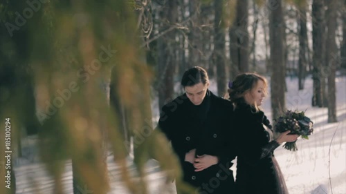 unusual wedding happy young lovers in black stand under tall pine tree. Gotic style. Beautiful brunette female in black coat, holding hand of man in black at sunny winter day. Slow Motion photo