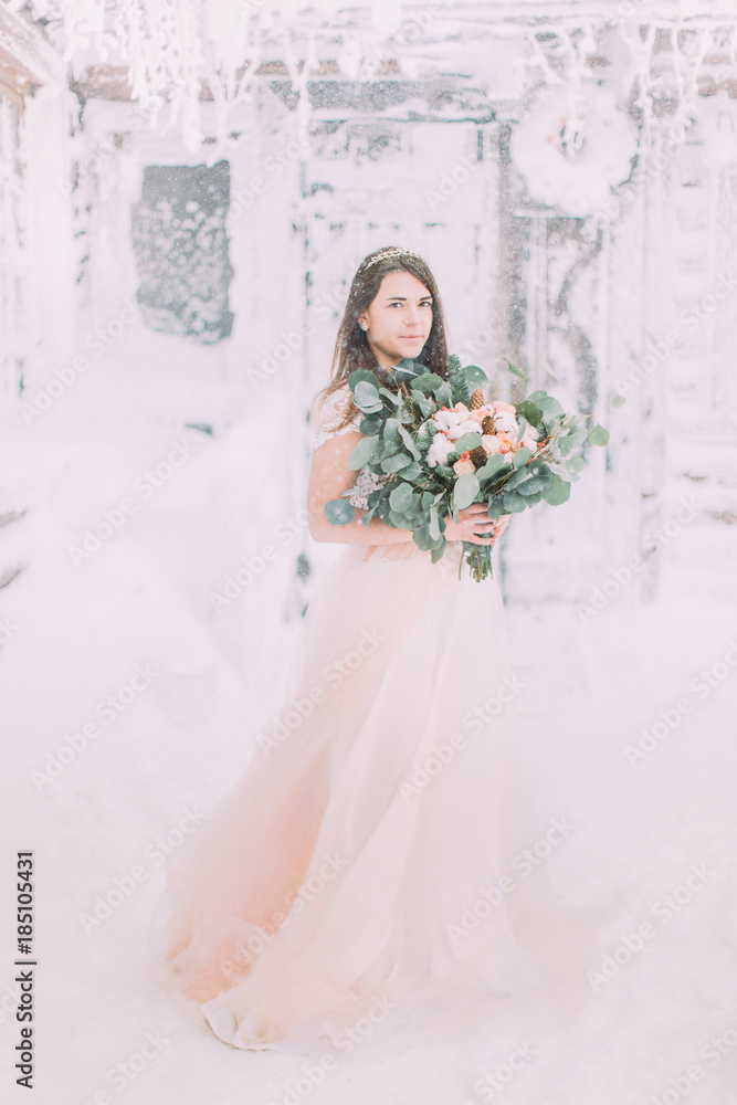 Bride in long pink dress holding wedding bouquet near the wooden house in winter mountains