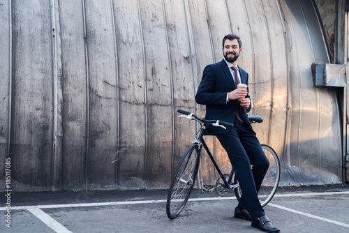 smiling businessman with disposable cup of coffee leaning on bicycle on street