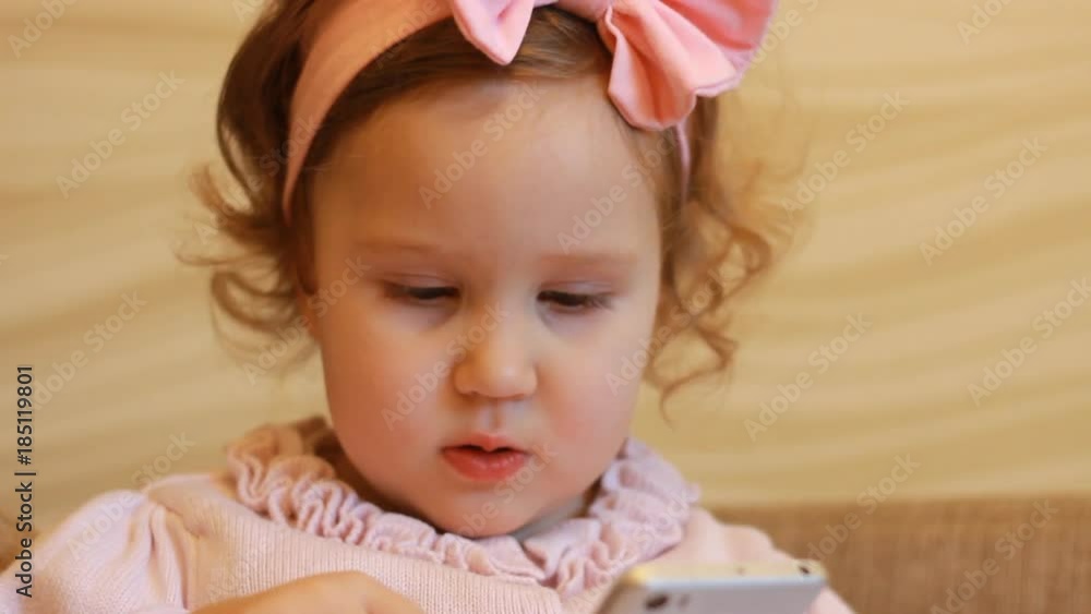 Funny child looks at the phone screen and plays downloaded application on a smart phone close-up. A little cute girl lies in sofa in a living room, looking cartoon and playing the game. Close-up. Funny child looks at the phone screen and plays downloaded application on a smart phone close-up. A little cute girl lies in sofa in a living room, looking cartoon and playing the game. Close-up.