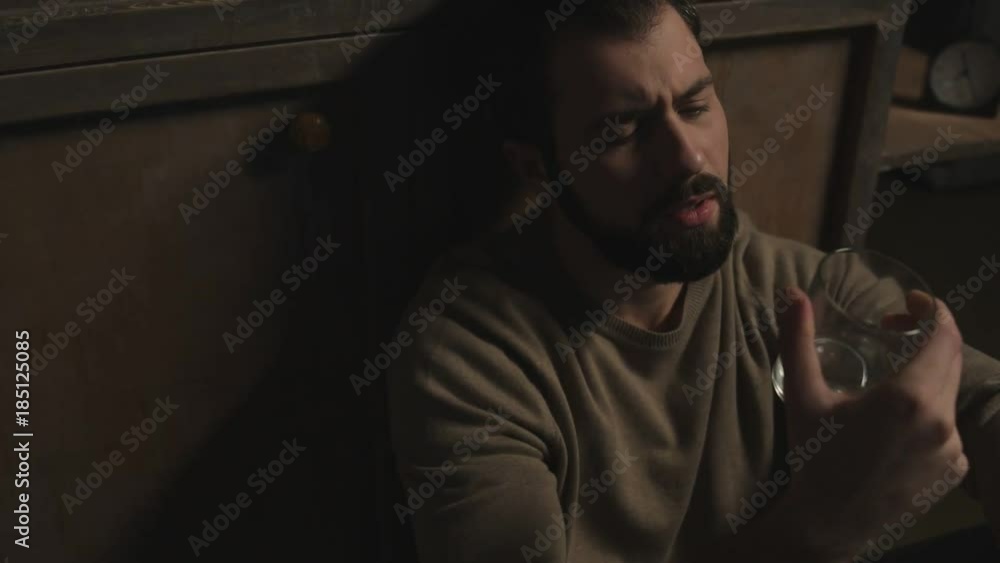 In despair. Close up of young bearded man sitting on the floor while drinking alcohol and expressing sadness In despair. Close up of young bearded man sitting on the floor while drinking alcohol and expressing sadness