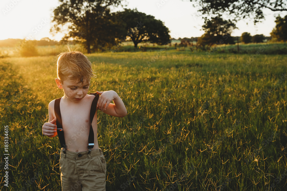 Shirtless boy wearing suspenders while standing amidst plants on field during sunset