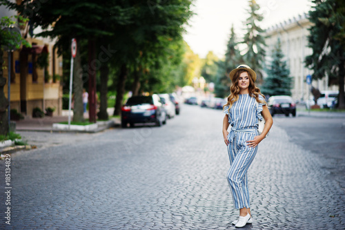 Portrait of a beautiful model in striped overall posing with hat and a backpack on a street with trees in a town. © AS Photo Family