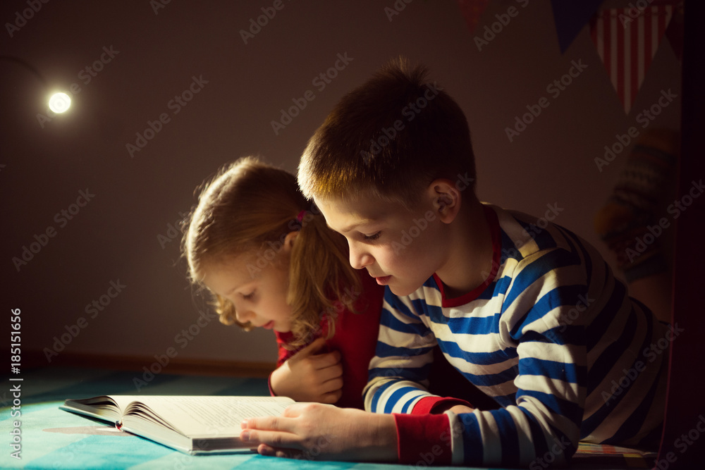 Two happy siblings reading book at darkness with flashlight at hiding place Two happy siblings reading book at darkness with flashlight at hiding place