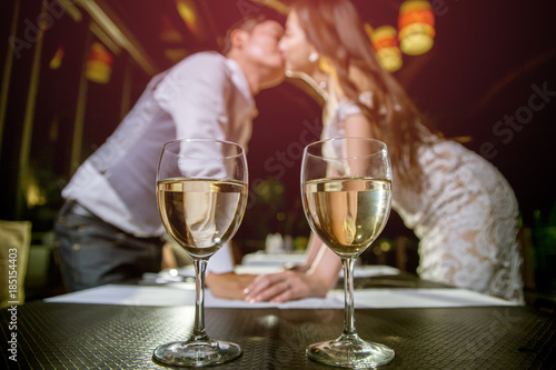 Two glasses of wine place on table. There are asian couple kissing together on blured background.