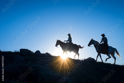 Silhouetted Western Cowboy and Cowgirl on horseback against a blue sky with sun flare at horizon photo