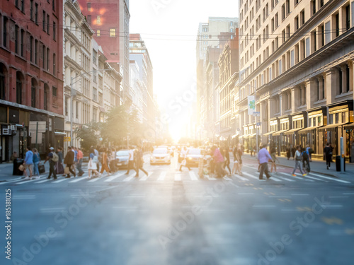 Crowd of anonymous people crossing the street at a busy intersection in Manhattan, New York City with the bright glow of sunset in the background © deberarr