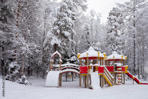 Snow-covered children's playground with slides and stairs in the park after a heavy snowfall against the background of snowy forest