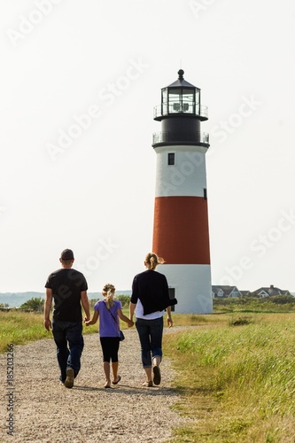 A family with mom, dad and young daughter walking up a path toward a light house on the east coast. Concept for travel in New England and the Atlanctic coast line. photo