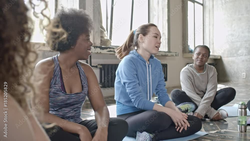PAN of young woman in fitness outfit sitting on mat and talking to cheerful female group before yoga class PAN of young woman in fitness outfit sitting on mat and talking to cheerful female group before yoga class