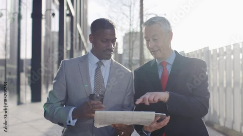 Two business professionals walk and talk over share prices in a paper, in slow motion