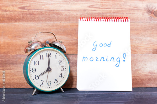 Vintage alarm clock and notebook with words Good morning on the wooden background.