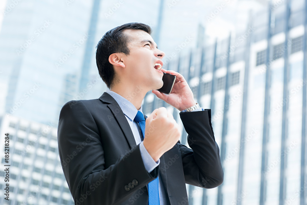 Winning businessman talking on mobile phone in the city, clenching his fist feeling great and delighted Winning businessman talking on mobile phone in the city, clenching his fist feeling great and delighted