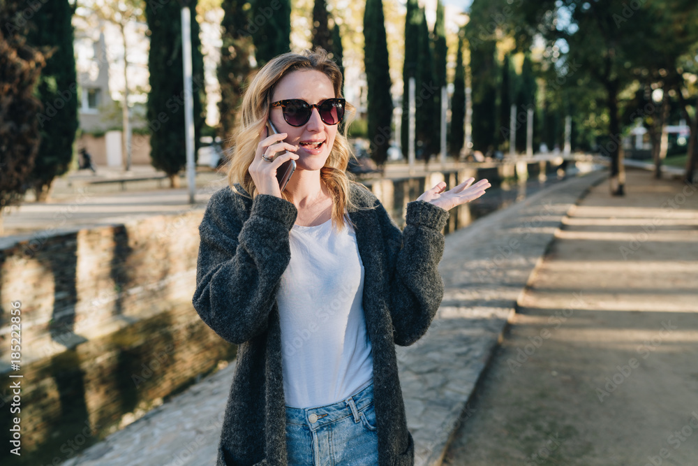 Fototapeta premium Summer evening. A young attractive woman in sunglasses stands in the park and talks happily on her cell phone. Telephone conversations. A girl is calling a friend on the phone. Lifestyle.