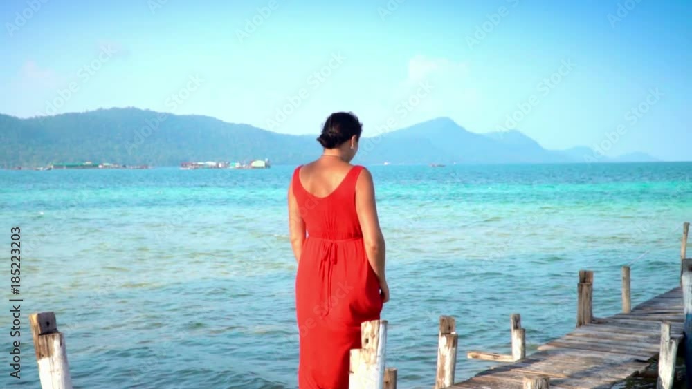 Beautiful woman walking down pier in long red dress. Phu Quoc Island. Landscape of mountains and sea. The woman walks on a pier on the island. Slow mothion Beautiful woman walking down pier in long red dress. Phu Quoc Island. Landscape of mountains and sea. The woman walks on a pier on the island. Slow mothion