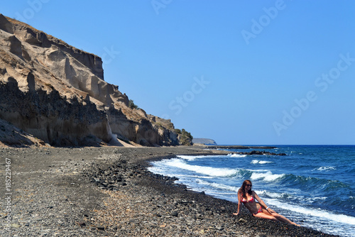 Young woman in sunglasses and in red swimsuit sits on dark gray sand/stones of Vourvoulos beach, Santorini, Greece. Background of blue sky and blue sea with waves.