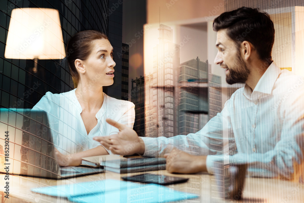 Unexpected details. Upbeat young man sitting at the table next to his female colleague and telling her news while the woman looking surprised Unexpected details. Upbeat young man sitting at the table next to his female colleague and telling her news while the woman looking surprised