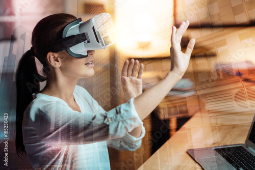 Brand new technology. Pleasant young woman sitting at the table in the office and testing a new VR headset, stretching out her hands as if being ready to touch something