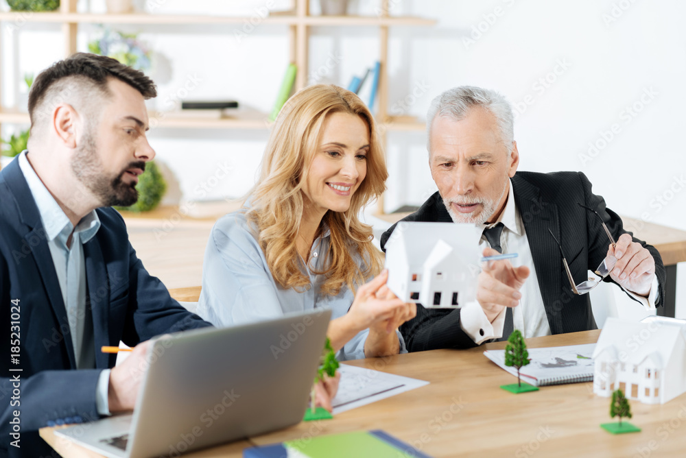 Amazing work. Smart interested aged real estate agent looking surprised and pointing at the lovely miniature house while his colleagues sitting by his side and looking attentively at it Amazing work. Smart interested aged real estate agent looking surprised and pointing at the lovely miniature house while his colleagues sitting by his side and looking attentively at it