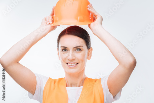 Orange hard hat. Attractive positive female worker  holding hard hat while standing on the isolated  background and grinning at the camera photo