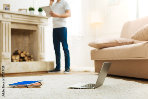 Modern technologies. Calm young man standing next to the big beautiful fireplace and holding an important notebook while his favorite modern laptop being placed on the floor next to the soft sofa