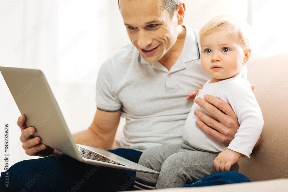 Calm child. Emotional cheerful young father sitting on a sofa with his child and smiling while looking at the screen of a convenient laptop Calm child. Emotional cheerful young father sitting on a sofa with his child and smiling while looking at the screen of a convenient laptop