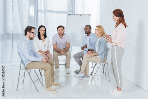 psychotherapist writing in notebook and talking with multiethnic people sitting on chairs during group therapy