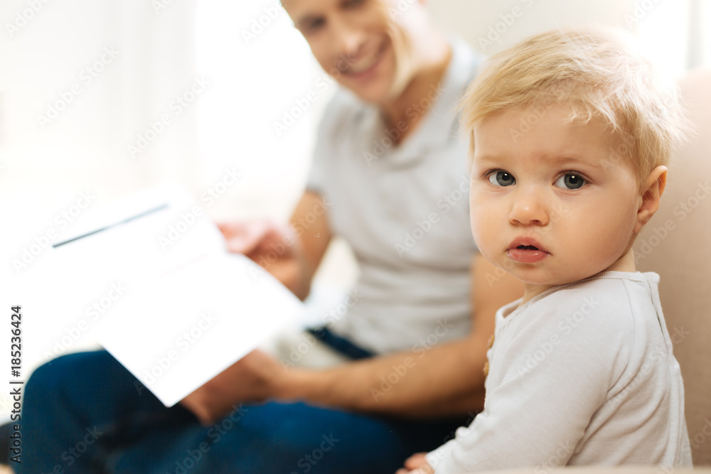 Big eyes. Pretty calm little child looking serious while being at home and sitting on the soft sofa together with a loving cheerful young father Big eyes. Pretty calm little child looking serious while being at home and sitting on the soft sofa together with a loving cheerful young father