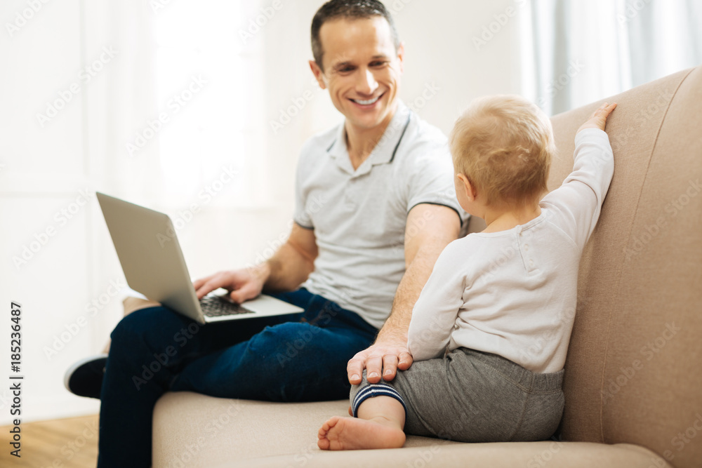 Loving parent. Handsome young attentive father sitting at home with a laptop on his knees and kindly looking at his little cute curious child and smiling Loving parent. Handsome young attentive father sitting at home with a laptop on his knees and kindly looking at his little cute curious child and smiling